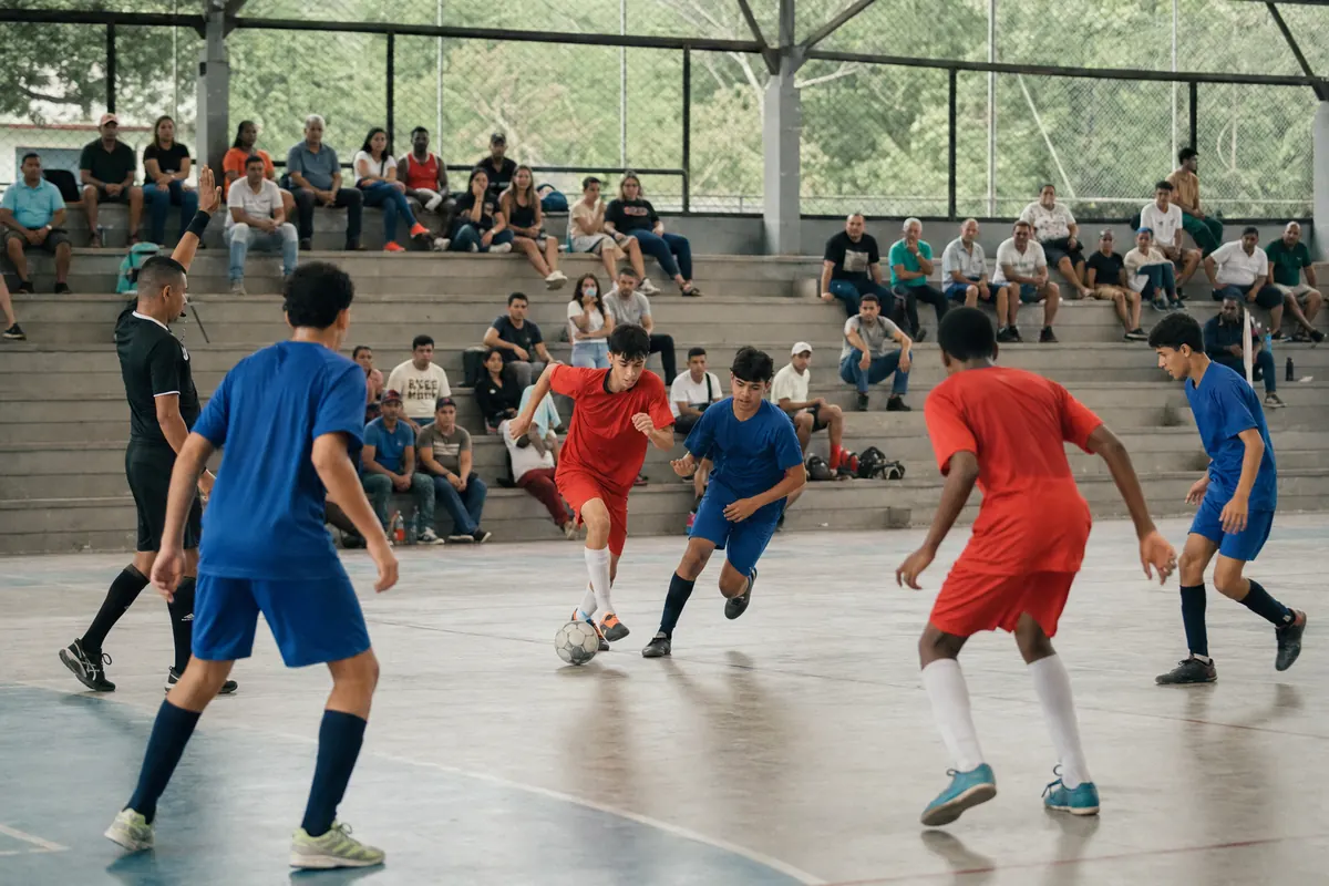 Youth futsal match in a municipal arena, with a referee and families in the stands