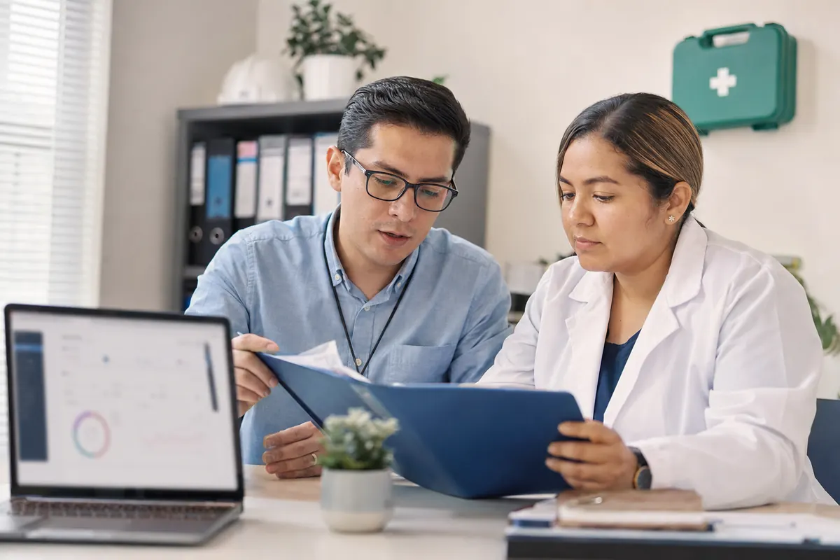 OHS professional and nurse review an occupational medical exam folder next to a laptop